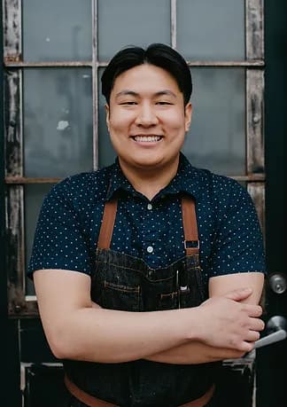 Chef Eric Pham, owner of Khue's Kitchen, standing with arms crossed and smiling in front of a rustic door.